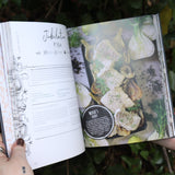 Someone holding the Natural Witch's Cookbook Hardcover Book open in front of a green bush, showing off the pages.