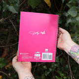 Someone holding the Dollyisms: Dolly Parton's 12-Month 2026 Softcover Planner in front of a green bush, showing off the back,