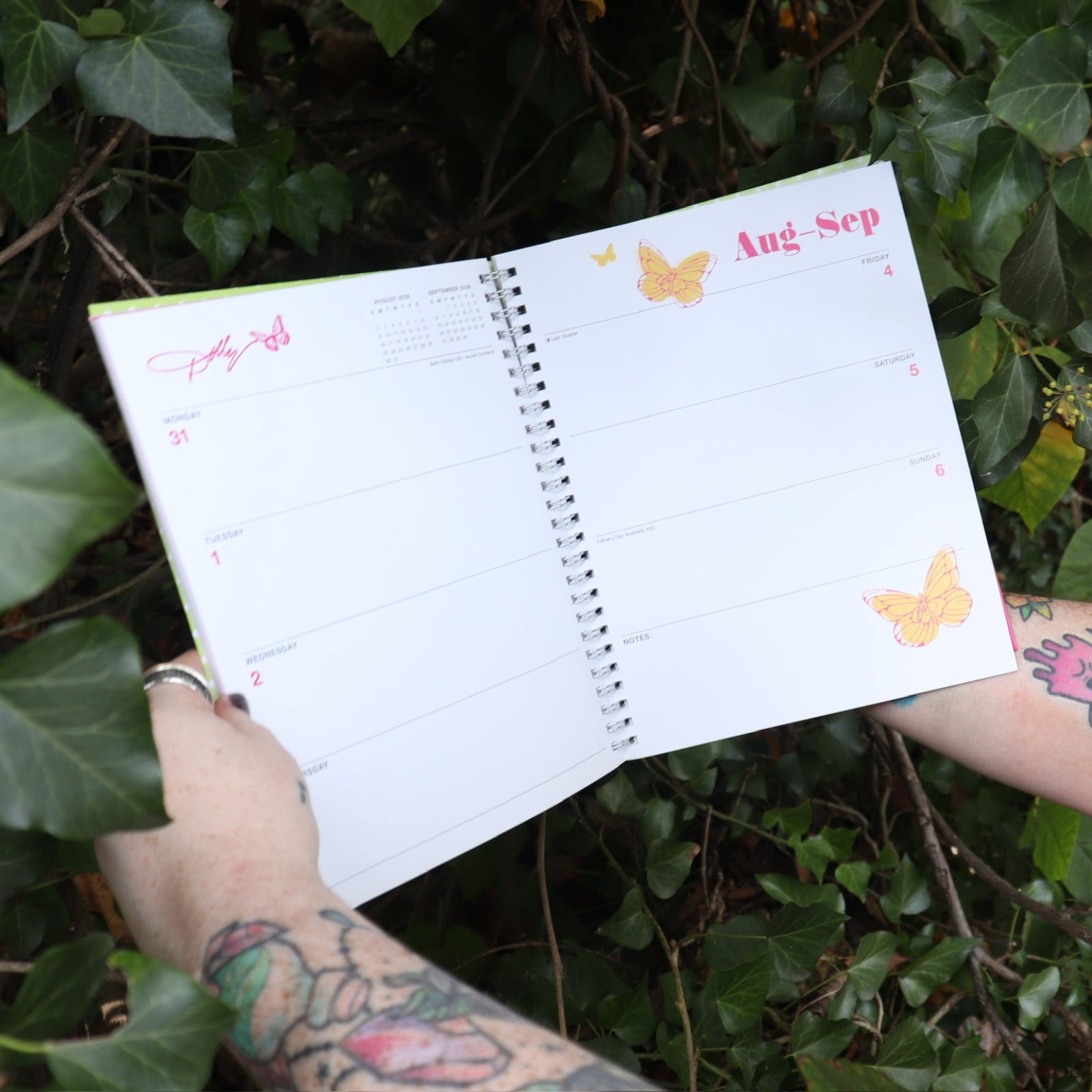 Someone holding the Dollyisms: Dolly Parton's 12-Month 2026 Softcover Planner open in front of a green bush, showing off the pages.