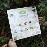 Someone holding the Kitchen Witch 2026 Wall Calendar in front of a green bush, showing off the back.