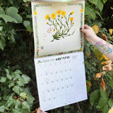 Someone holding the Kitchen Witch 2026 Wall Calendar open in front of a green bush.