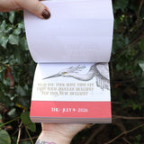 Someone holding the Effin' Birds 2026 Day-to-Day Calendar open in front of a green bush, showing off the pages.