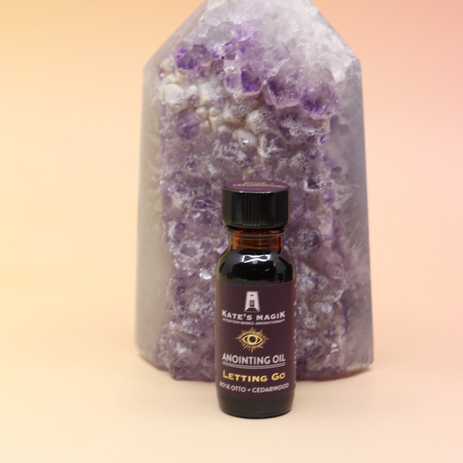The Letting Go Reiki Charged Anointing Oil in front of an amethyst tower all on a pink and yellow background.