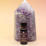 The Sun of Success Reiki Charged Anointing Oil in front of an amethyst tower all on a pink and yellow background.