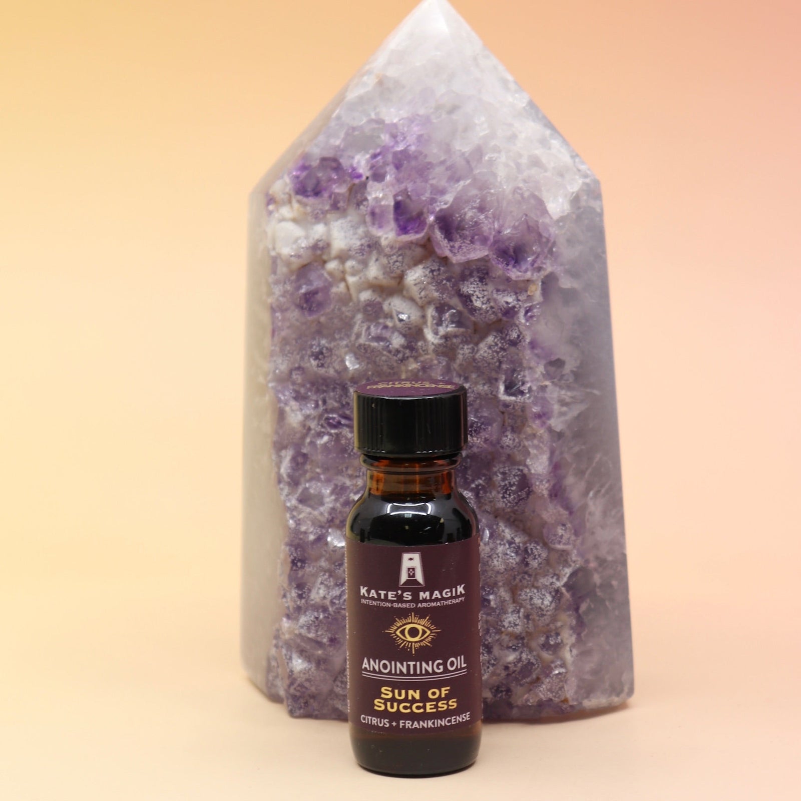 The Sun of Success Reiki Charged Anointing Oil in front of an amethyst tower all on a pink and yellow background.