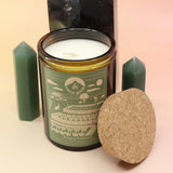 The Deeply Rooted 12oz Soy Candle in front of a large crystal tower surrounded by two smaller crystal towers all on a pink and yellow background.