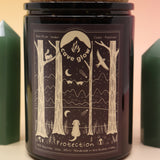 The Protection 12oz Soy Candle in front of a large crystal tower surrounded by two smaller crystals all on a pink and yellow background.