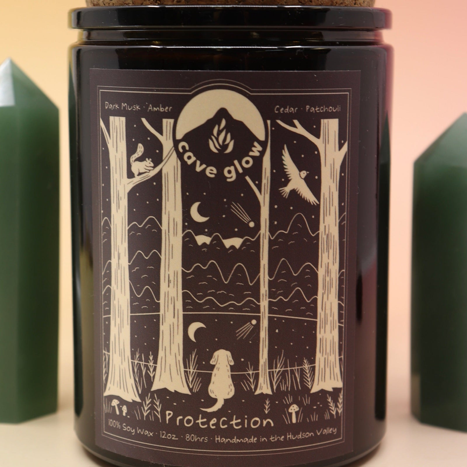 The Protection 12oz Soy Candle in front of a large crystal tower surrounded by two smaller crystals all on a pink and yellow background.