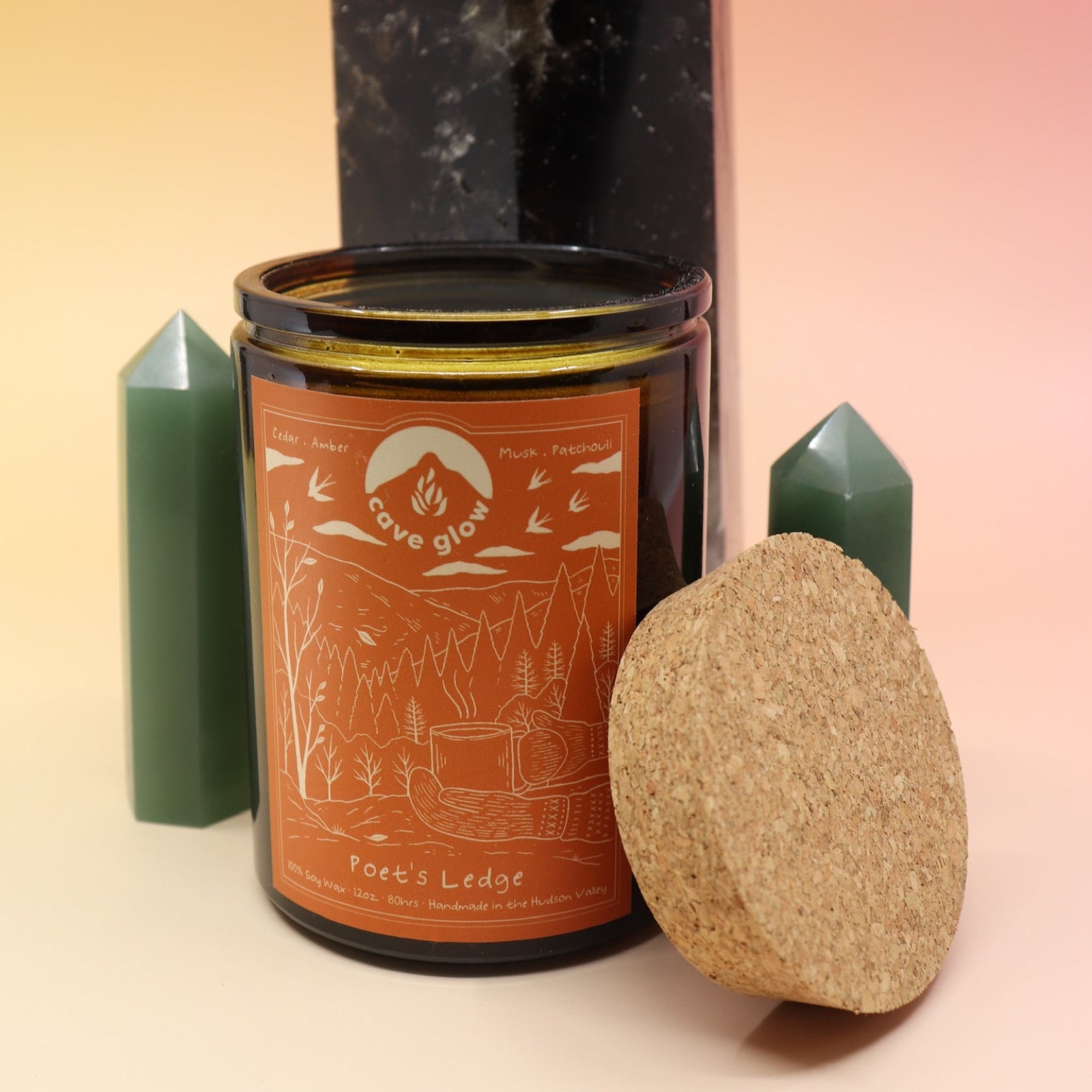 The Poet's Ledge 12oz Soy Candle in front of a large crystal tower and surrounded by two smaller crystal towers all on a pink and yellow background.