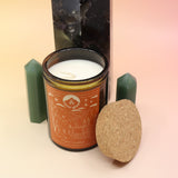 The Poet's Ledge 12oz Soy Candle in front of a large crystal tower and surrounded by two smaller crystal towers all on a pink and yellow background.