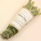 The Piñon Pine Sacred Smoke Cleansing Stick with Quartz Crystal on a pink and yellow background.