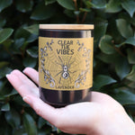 Someone holding the Clear the Vibes Soy Wax Garden Energy Clearing Candle in front of a green bush.