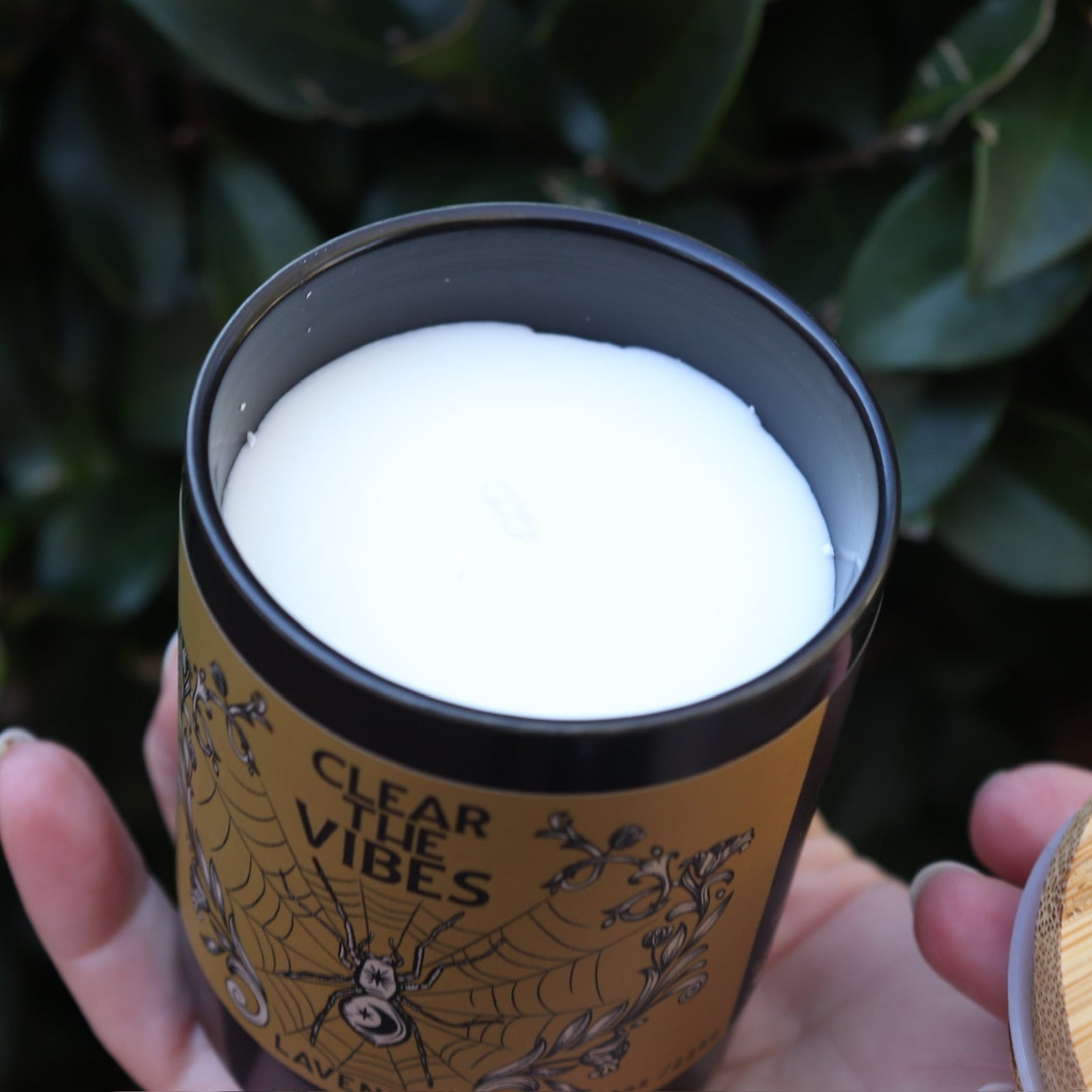 Someone holding the Clear the Vibes Soy Wax Garden Energy Clearing Candle in front of a green bush, showing off the inside.