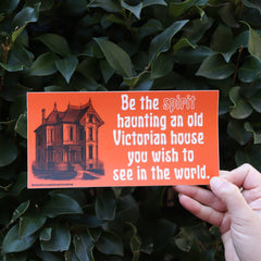 Someone holding the Be the Spirit Haunting a Victorian Halloween Bumper Sticker in front of a green bush.