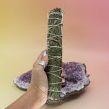 The White Sage & Cedar Smudge Stick 9 in. being held in front of an amethyst piece all on a pink and yellow background.