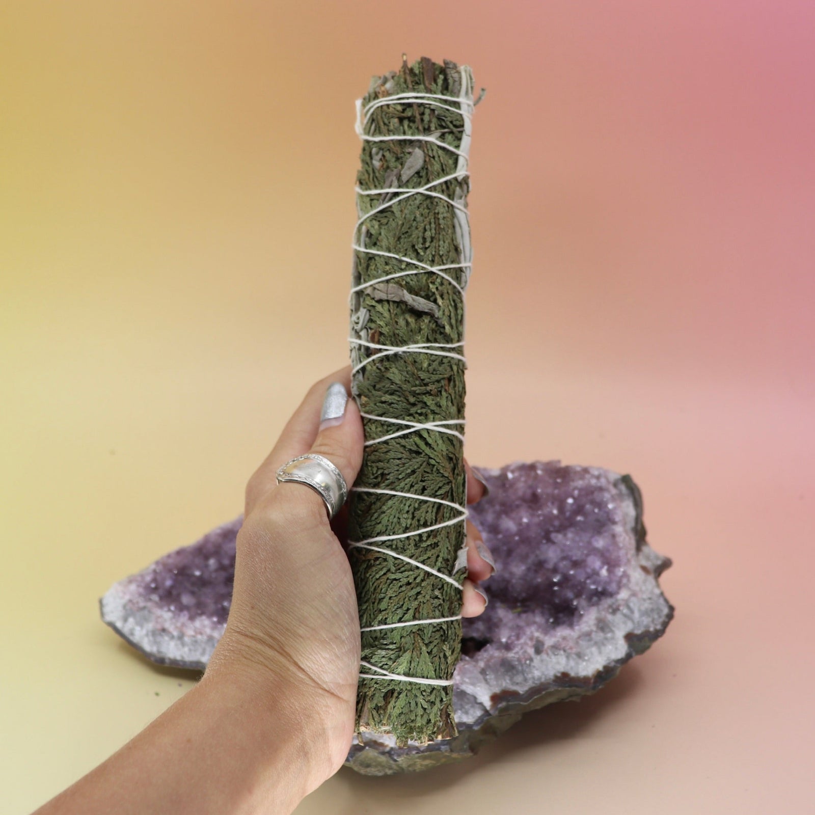 The White Sage & Cedar Smudge Stick 9 in. being held in front of an amethyst piece all on a pink and yellow background.