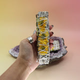 The Sunflower White Sage Smudge Stick 6 in. being held in front of another Sunflower White Sage Smudge Stick 6 in. on an amethyst piece all on a pink and yellow background.