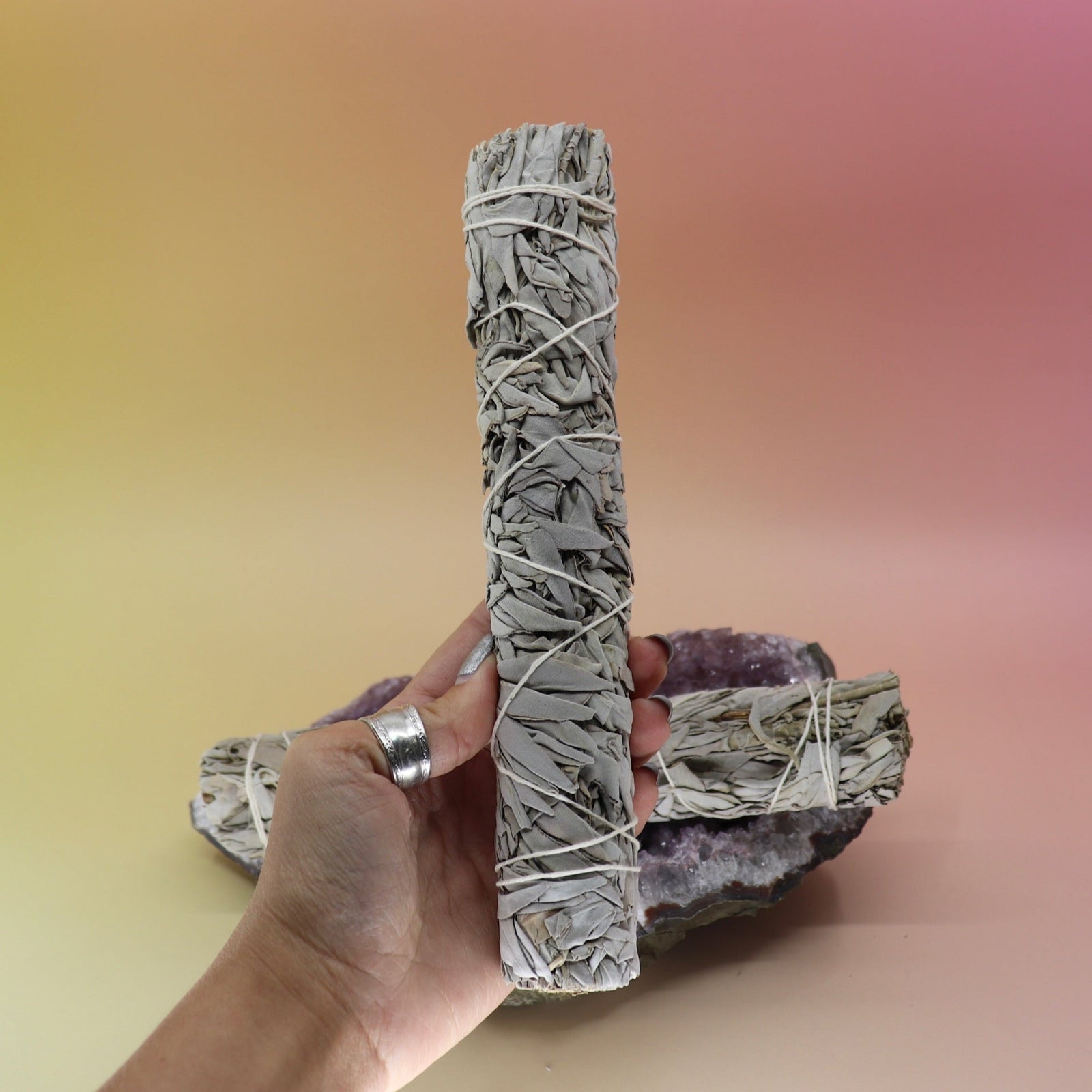 Someone holding the White Sage 9 in. Smudge Stick for Cleansing in front of an amethyst piece all on a pink and yellow background.