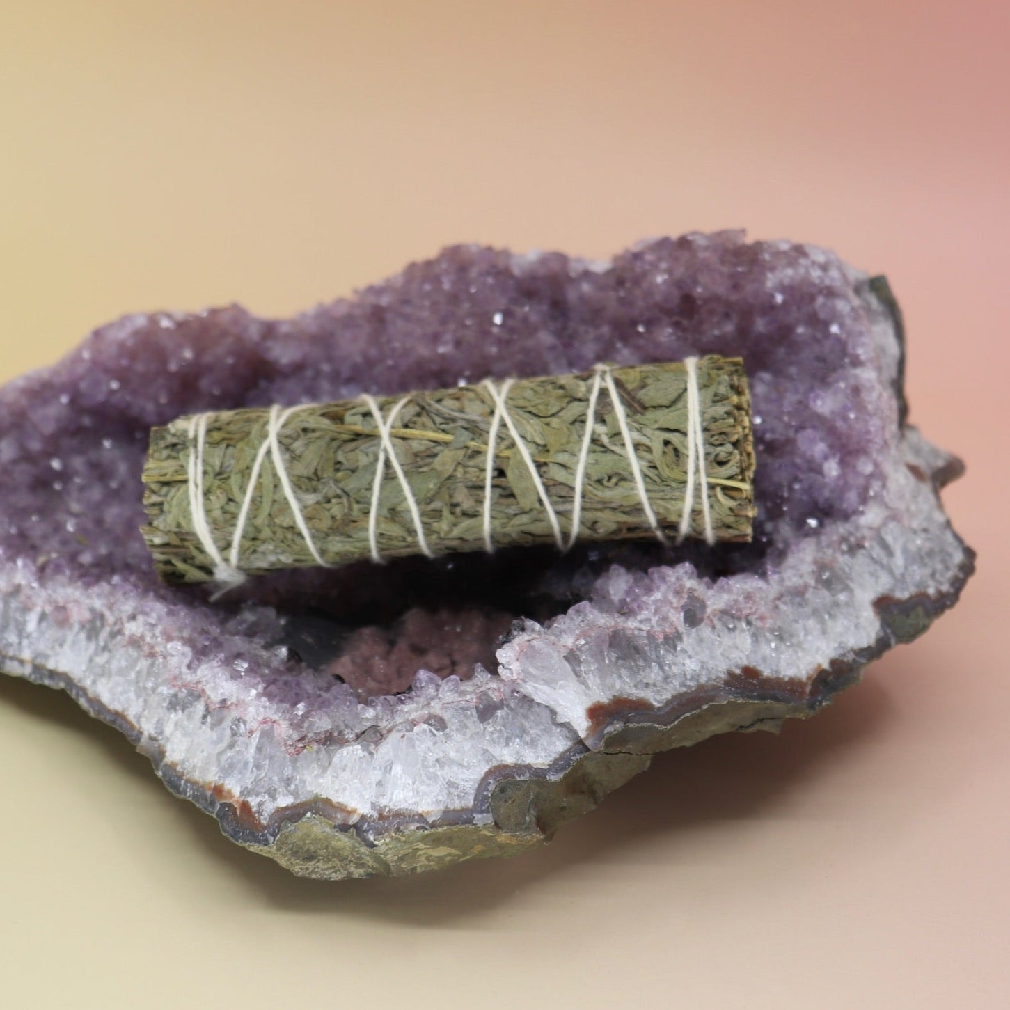 The Organic Mugwort Sage - 4 in. Smudge Stick on an amethyst piece all on a pink and yellow background.