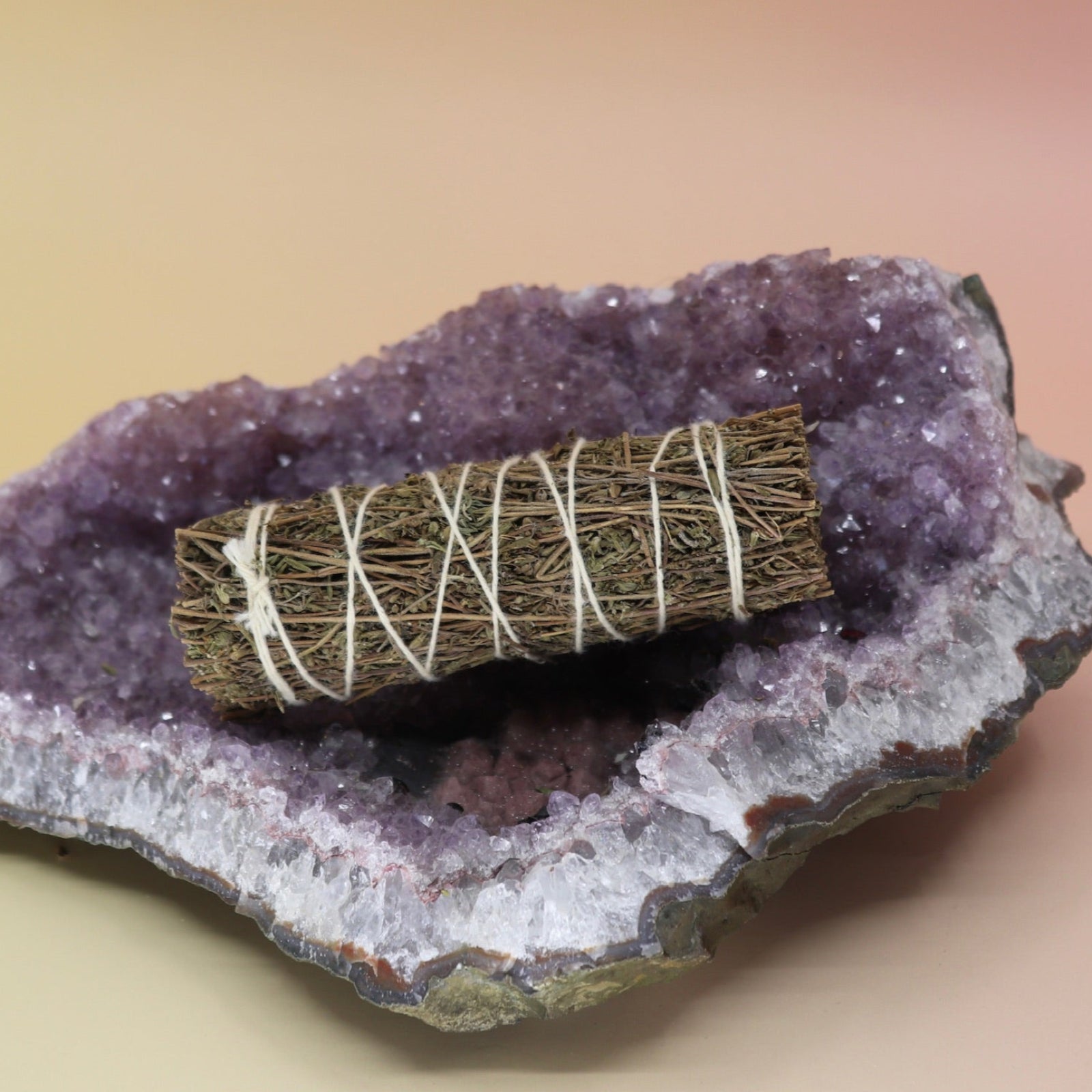 The Organic Thyme Sage - 4 in. Smudge Stick on an amethyst piece all on a pink and yellow background.