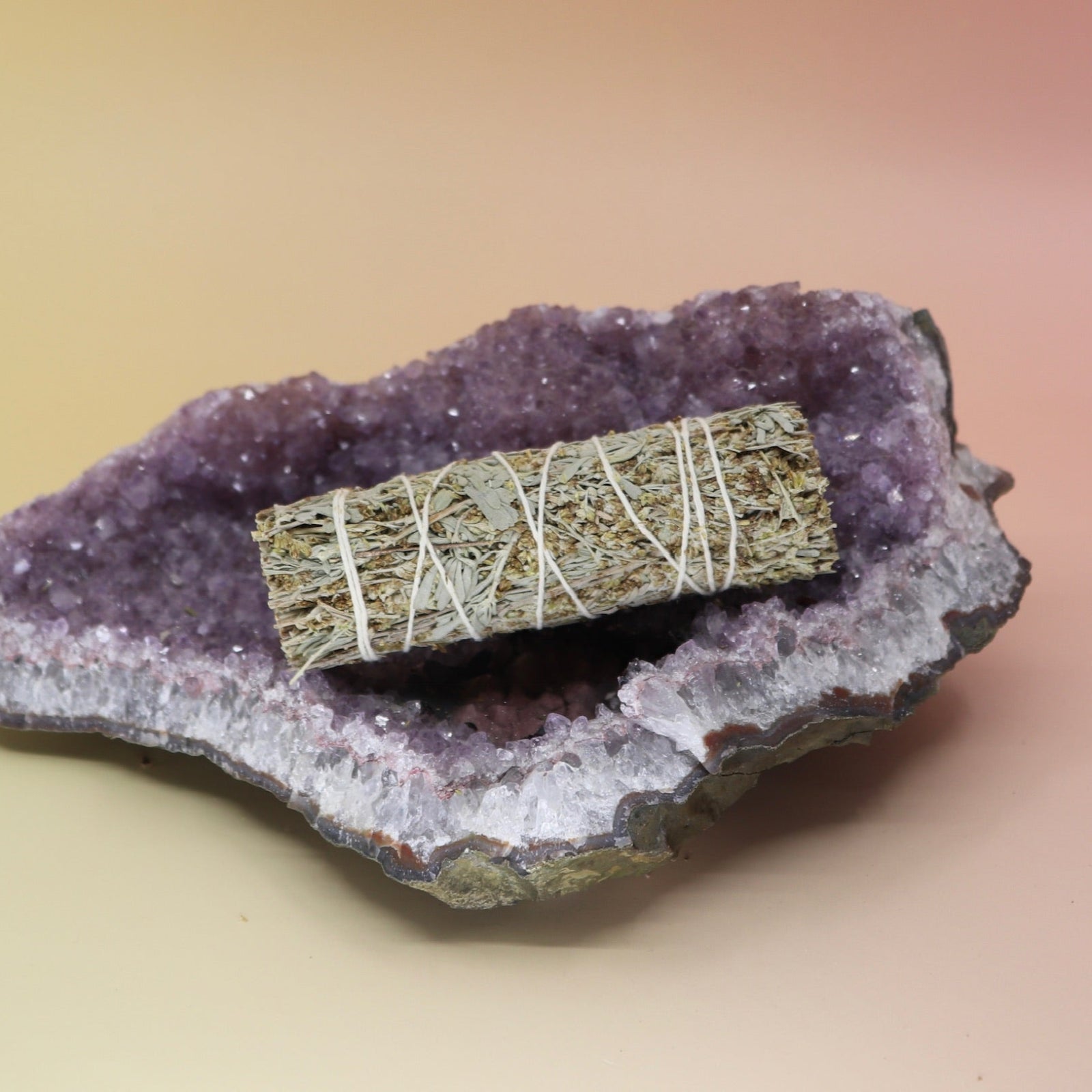 The Organic Blue Sage - 4 in. Smudge Stick on an amethyst piece all on a pink and yellow background.