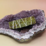 The Organic Juniper Sage - Protection and Harmony 4 in. Smudge Stick on an amethyst piece all on a pink and yellow background.