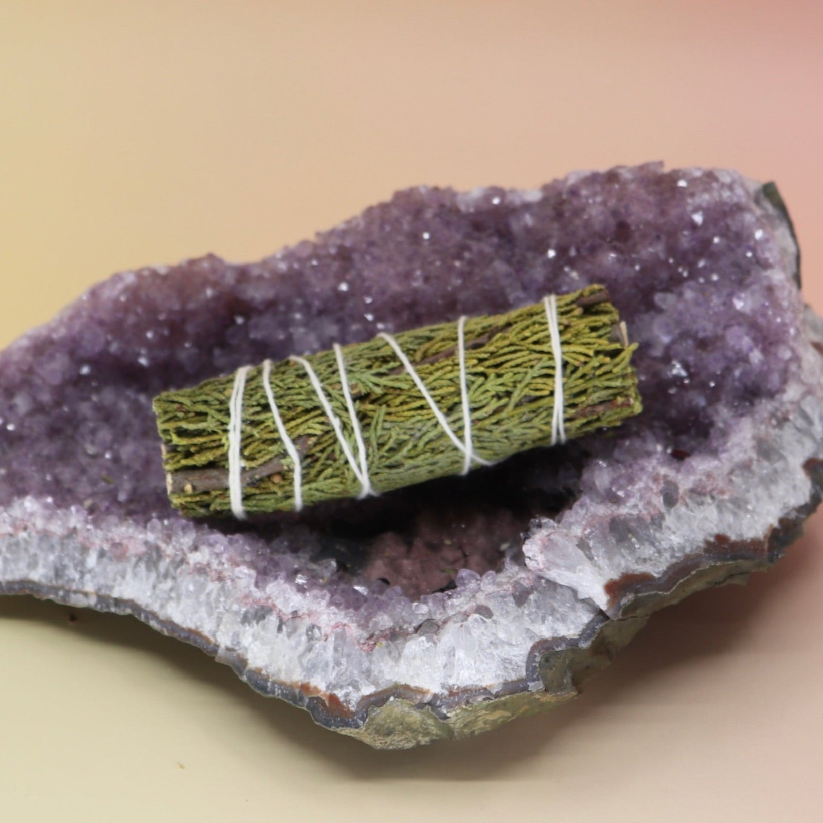 The Organic Juniper Sage - Protection and Harmony 4 in. Smudge Stick on an amethyst piece all on a pink and yellow background.