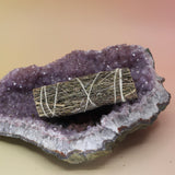 The Organic Lavender Sage - 4 in. Smudge Stick on an amethyst piece all on a pink and yellow background.