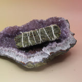 The White Sage & Mugwort Smudge Stick 4 in. on an amethyst piece on a pink and yellow background.