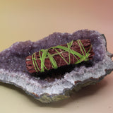 The Secret Garden Floral Dragons Blood Sage - 4 in. Smudge Stick on an amethyst piece all on a pink and yellow background.
