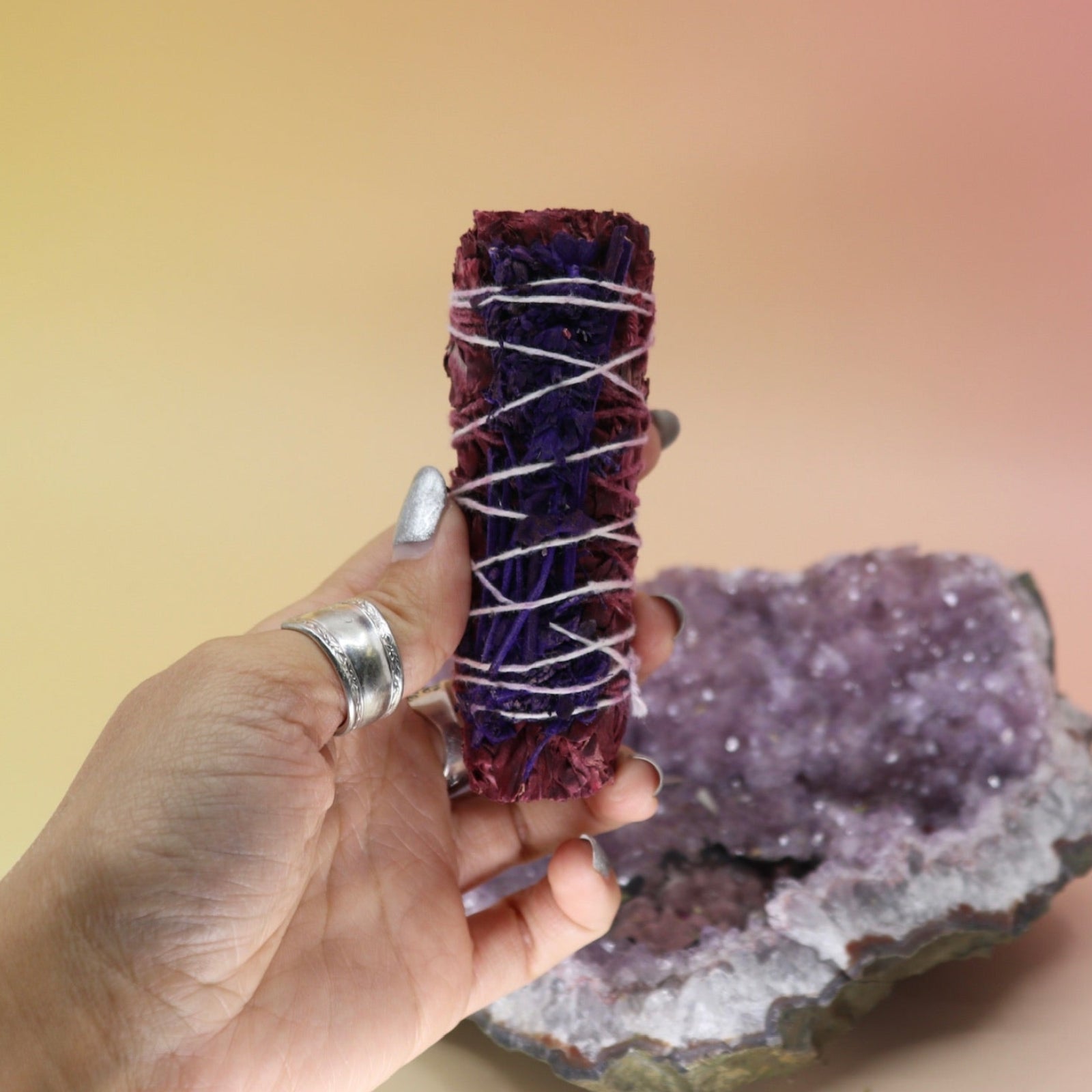 Someone holding the Dragon Blood Sage with Lavender - 4 in. Smudge Stick in front of an amethyst piece on a pink and yellow background.