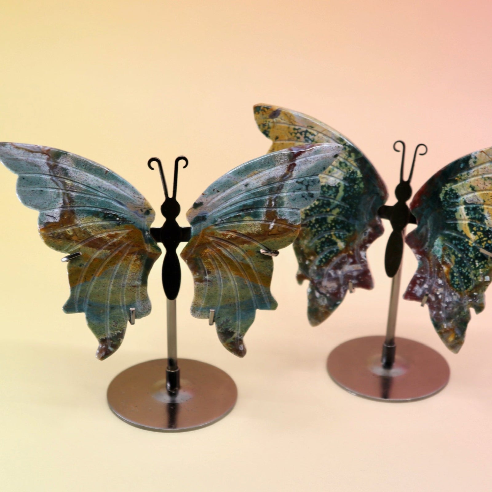 Two of the Natural Marine Jasper Butterfly Crystals with Stand on a pink and yellow background.