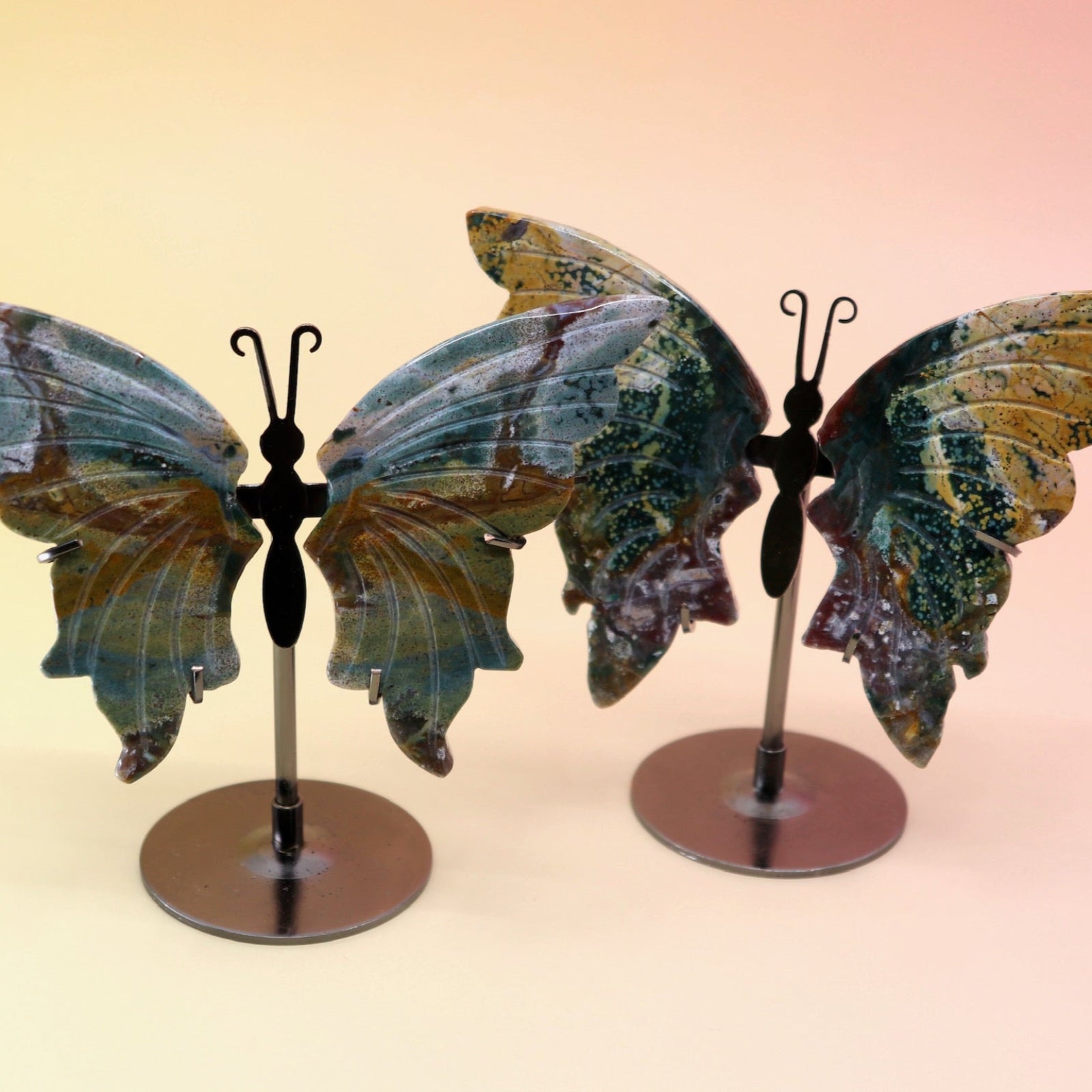 Two of the Natural Marine Jasper Butterfly Crystals with Stand on a pink and yellow background.