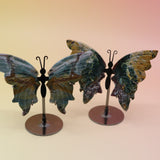 Two of the Natural Marine Jasper Butterfly Crystals with Stand on a pink and yellow background.