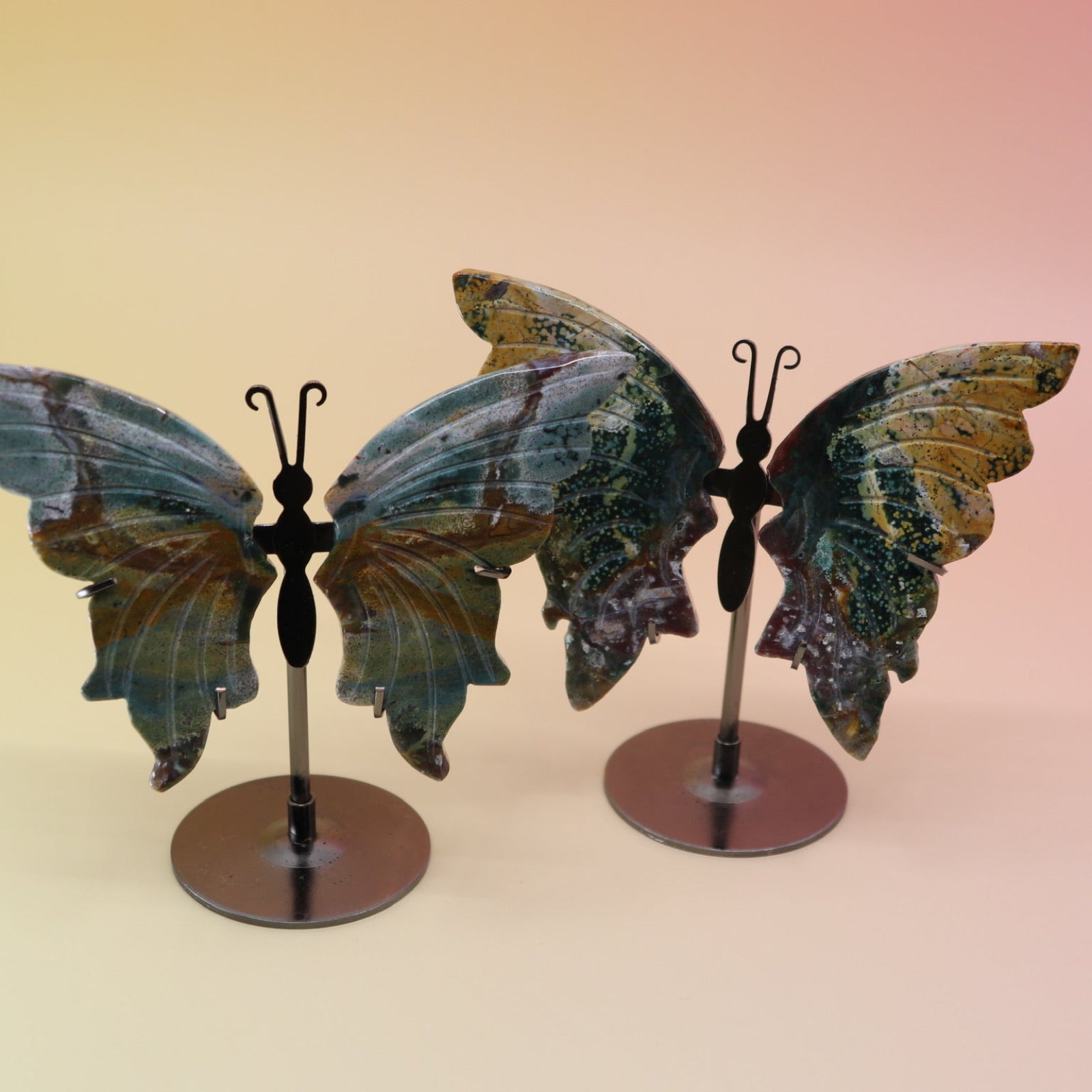 Two of the Natural Marine Jasper Butterfly Crystals with Stand on a pink and yellow background.