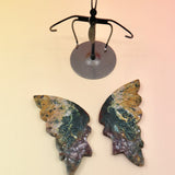 Two of the Natural Marine Jasper Butterfly Crystal with Stand on a pink and yellow background with the crystal butterfly wings off the stand.