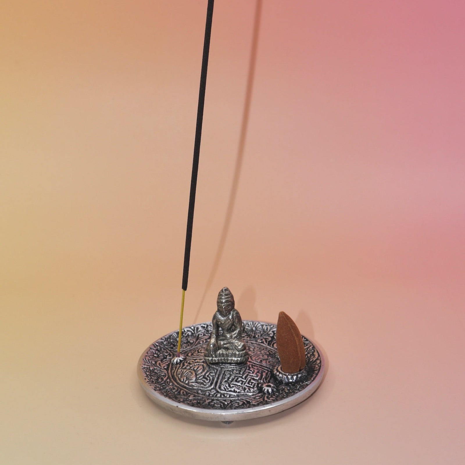 The Silver Buddha Cone and Stick Incense Holder on a pink and yellow background an incense cone and an incense stick on it.