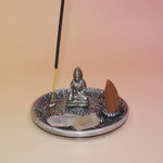 The Silver Buddha Cone and Stick Incense Holder on a pink and yellow background with crystals, an incense cone, and an incense stick on it.