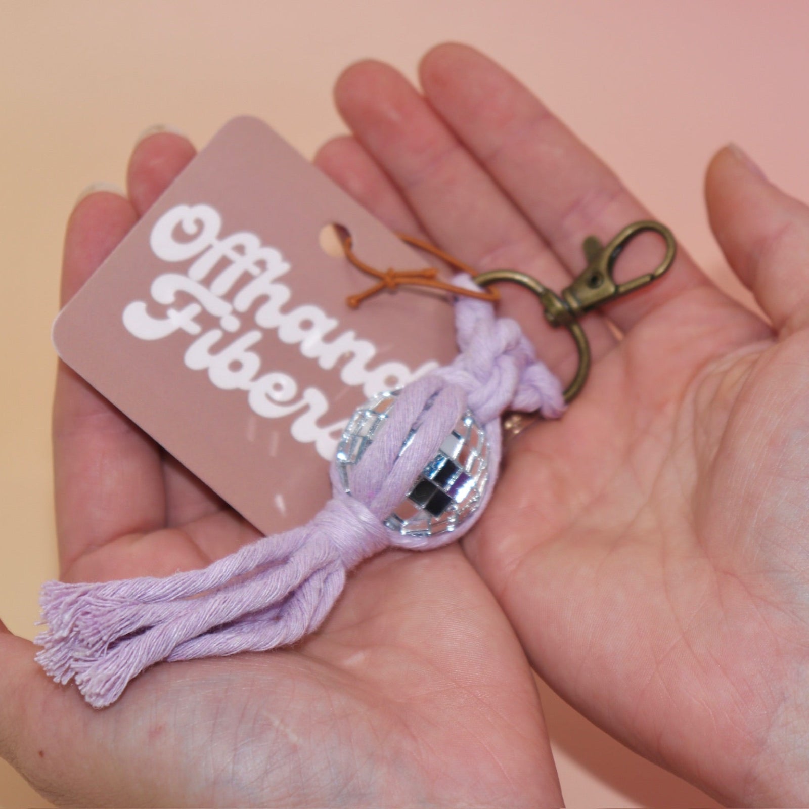 Someone holding the lavender Mini Macrame Disco Ball Keychain in front of a pink background.