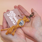 Someone holding the mustard Mini Macrame Disco Ball Keychain in front of a pink background.