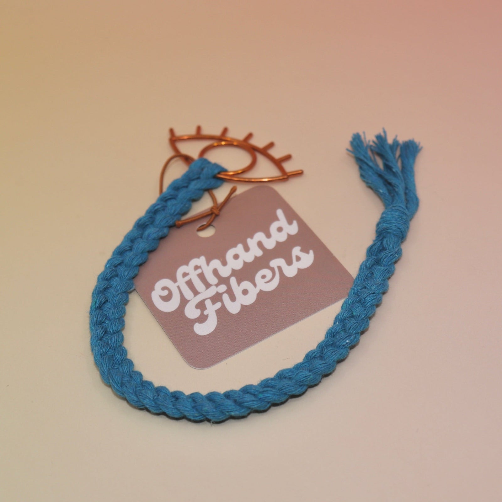 The peacock blue All Seeing Eye Charm Macrame 12 in. Bookmark on a light pink and yellow background.