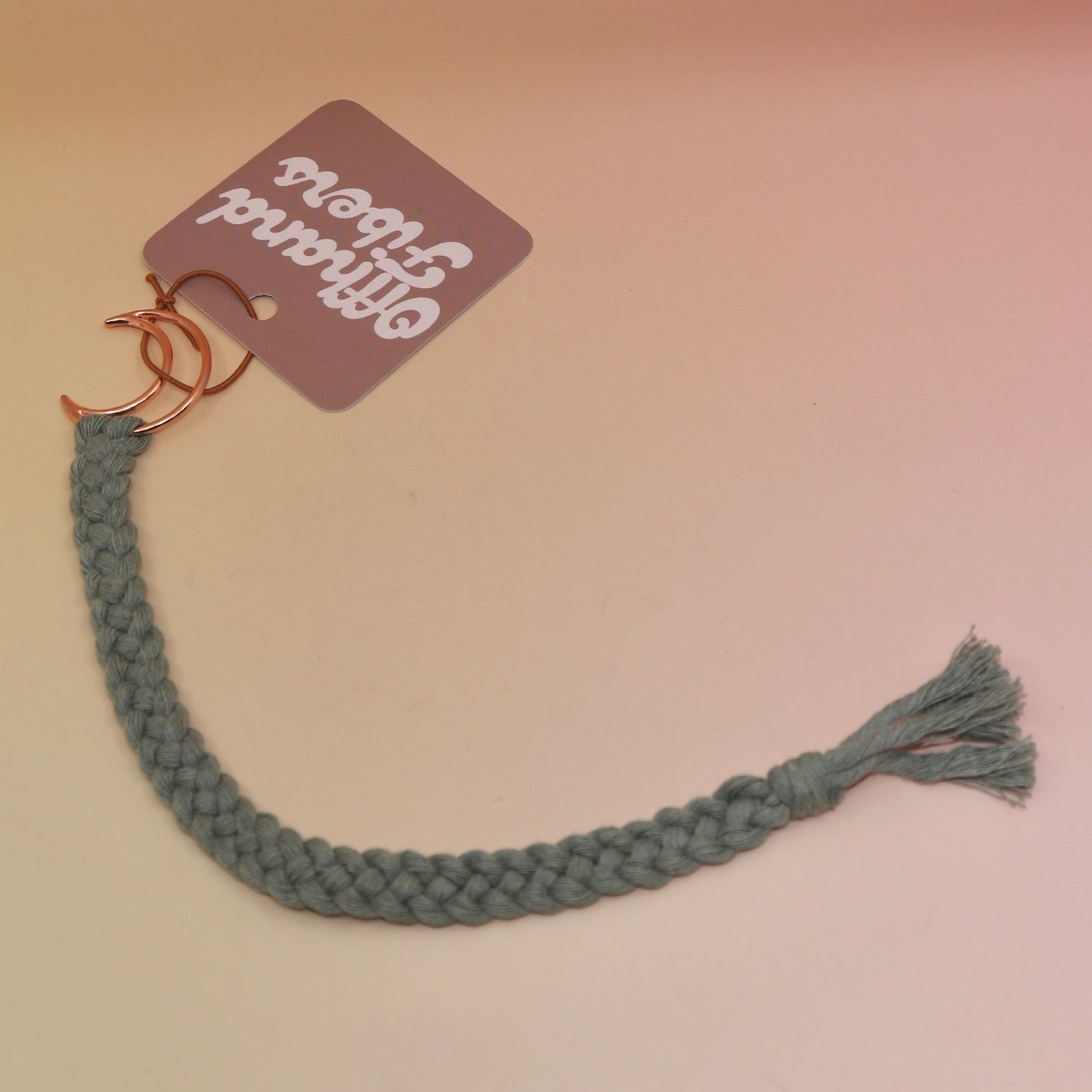 The sage Boho Moon Charm Macrame 12 in. Bookmark on a light pink and yellow background.