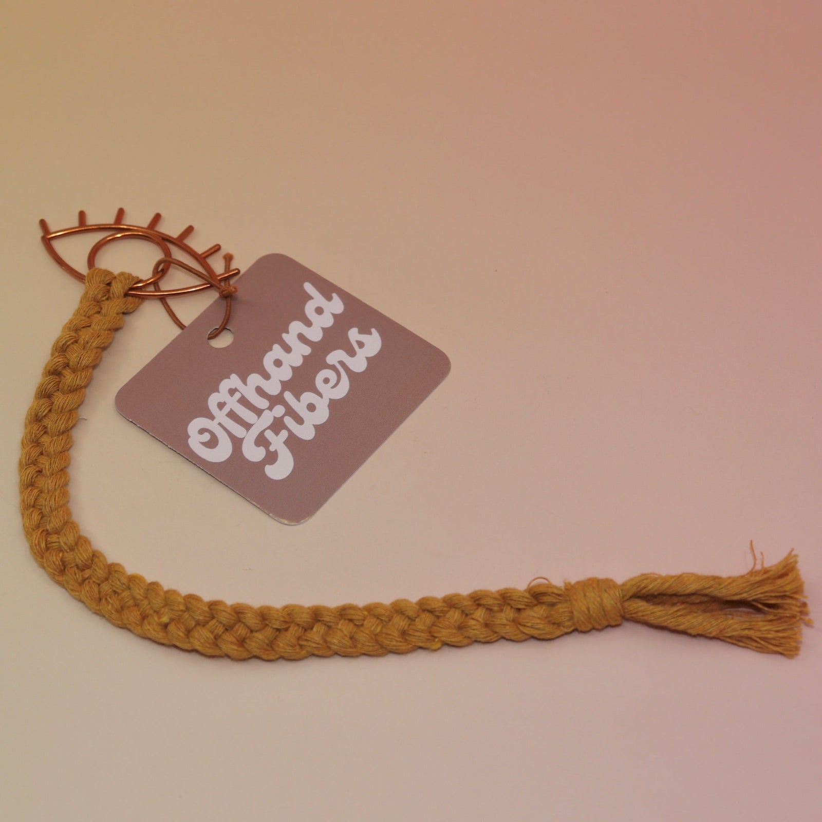 The mustard All Seeing Eye Charm Macrame 12 in. Bookmark on a light pink and yellow background.