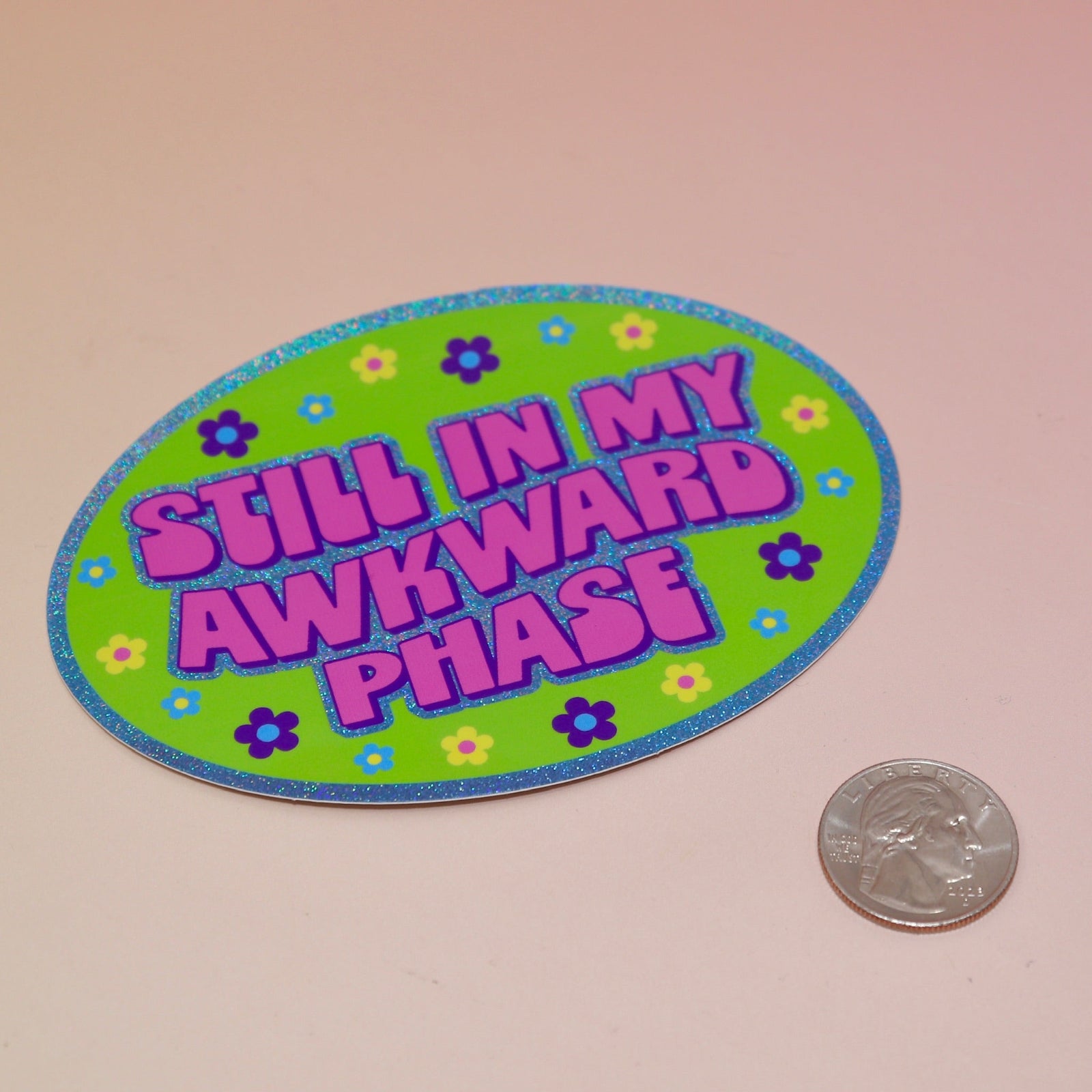 The Still In My Awkward Phase Oval Holographic Glitter Sticker on a light pink background, next to a quarter.