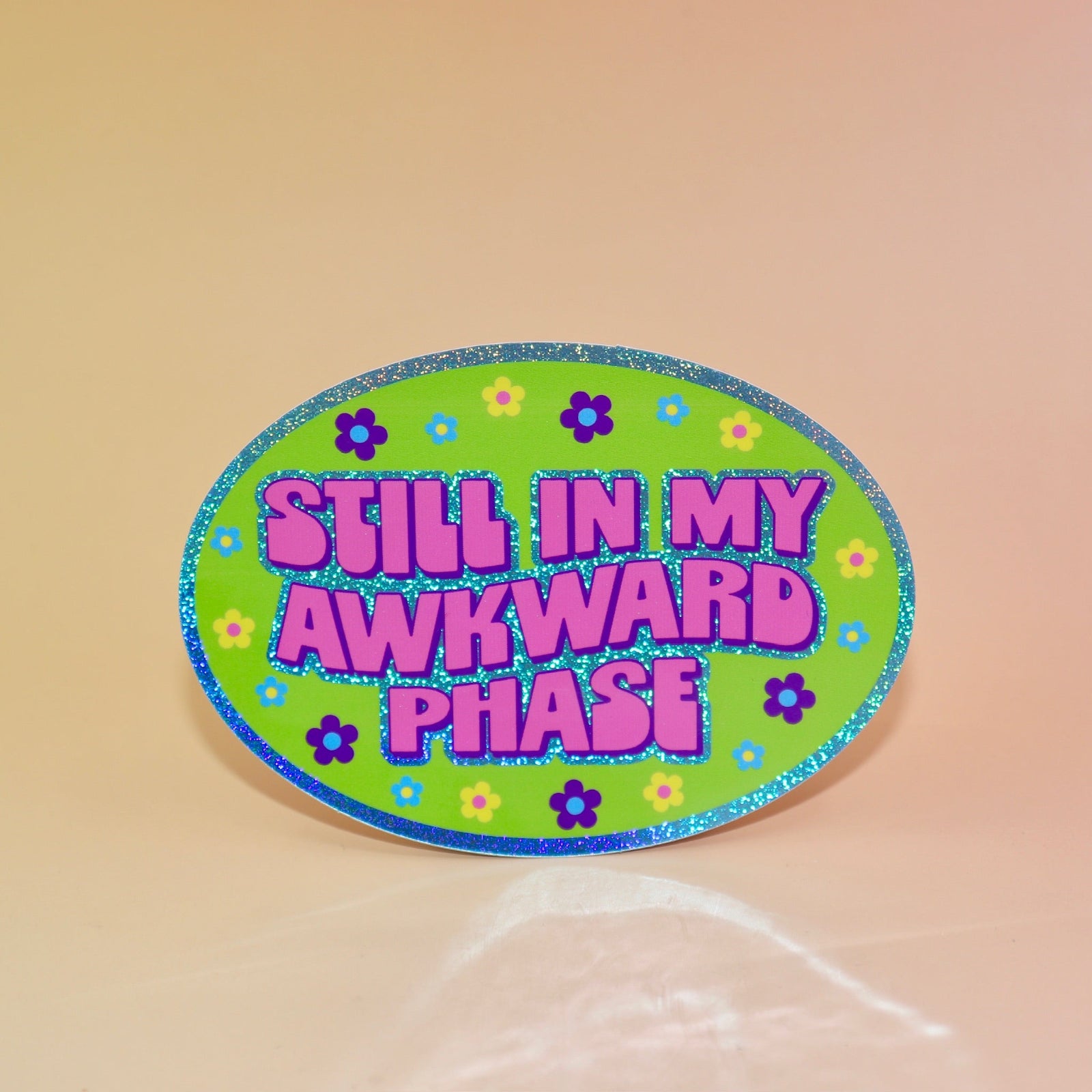 The Still In My Awkward Phase Oval Holographic Glitter Sticker on a light pink background.