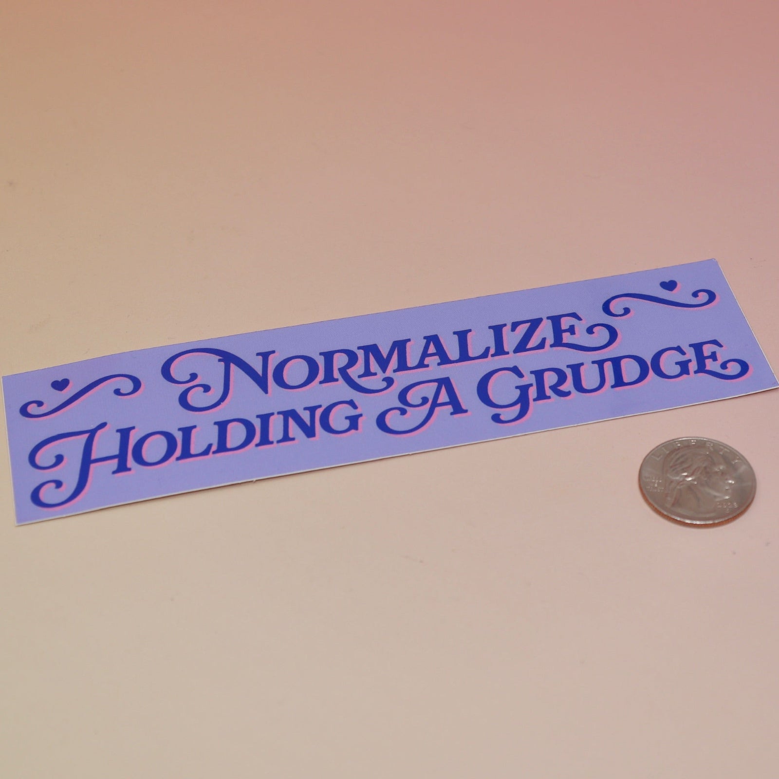 The Normalize Holding A Grudge Vinyl Bumper Sticker on a pink and yellow background, next to a quarter.