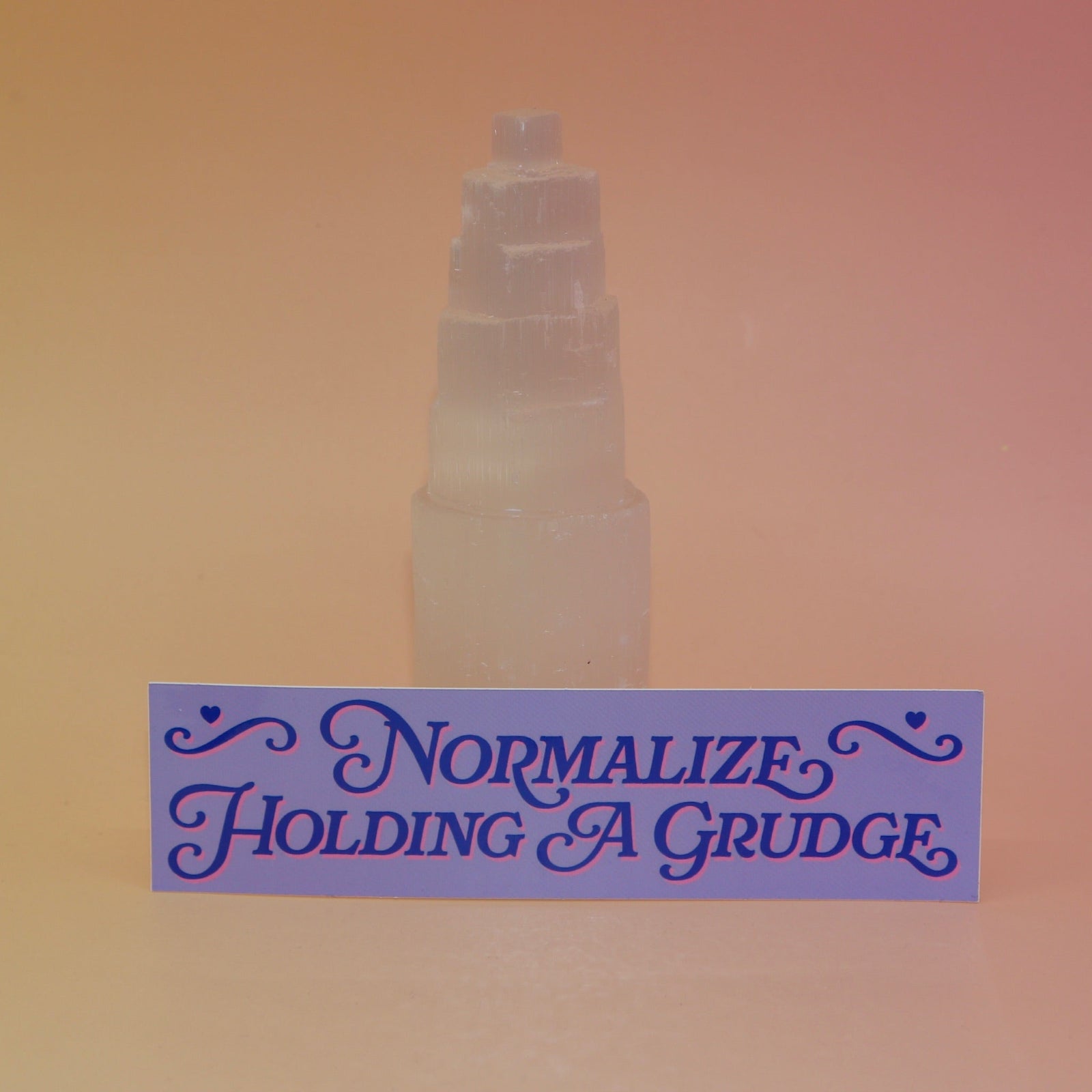 The Normalize Holding A Grudge Vinyl Bumper Sticker against a selenite tower on a pink and yellow background.