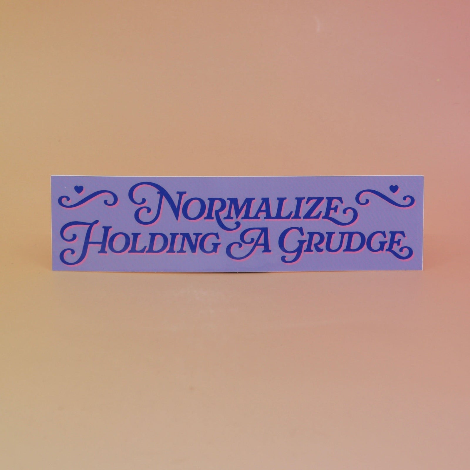 The Normalize Holding A Grudge Vinyl Bumper Sticker on a pink and yellow background.