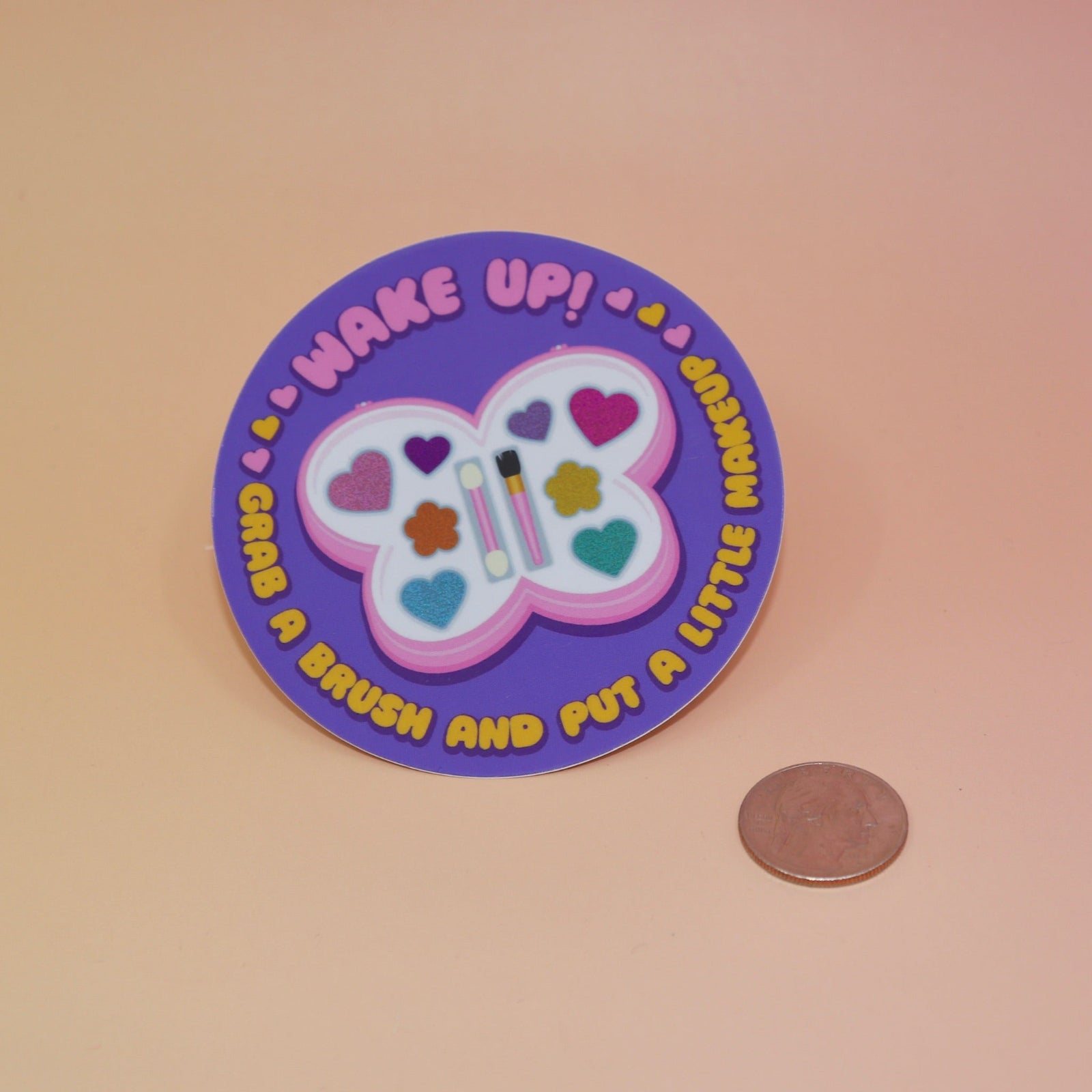 The Wake Up Grab A Brush Makeup SOAD Holographic Glitter Vinyl Sticker next to a quarter on a light pink background.
