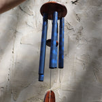 The Woodstock Dahlia Garden Wind Chime in front of a cement wall.